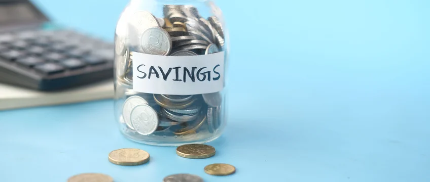 Glass savings jar filled with coins next to a calculator on a blue background