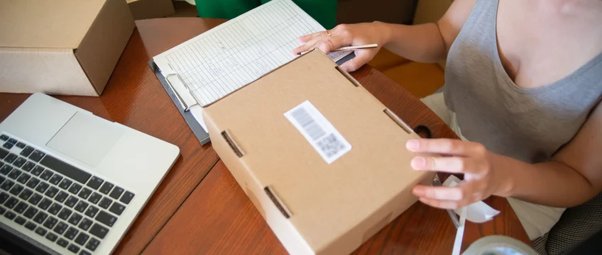 Person packing a parcel on a desk beside a laptop, preparing an online order for shipping